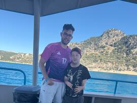 Zwei Personen stehen auf einem Boot vor Meer und felsiger Küste. Im Hintergrund sind Berge und ein heller Strand zu sehen.