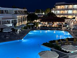 Eine Hotelanlage mit beleuchtetem Pool, Liegen und Sonnenschirmen ist am Abend zu sehen. Im Hintergrund befinden sich helle Gebäude und ein Restaurantbereich.