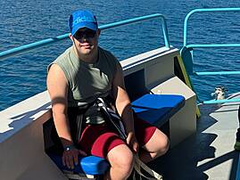 Eine Person sitzt auf einem Boot vor dem blauen Meer. Im Hintergrund ist eine felsige Küste zu sehen.