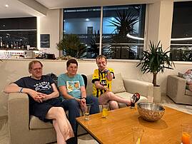 Drei Personen sitzen abends in einer Hotellounge auf einem Sofa. Vor ihnen stehen Getränke auf einem Tisch.