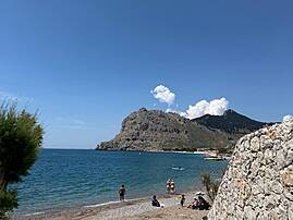 Ein Strand auf Rhodos liegt vor türkisblauem Meer und felsigen Bergen. Mehrere Menschen entspannen am Wasser.