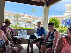 Eine Reisegruppe sitzt im Schatten an einem Tisch nahe dem Pool. Im Hintergrund sind Palmen, Hotelgebäude und Liegen zu sehen.