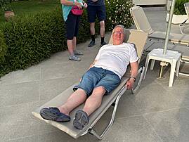 Drei Personen halten sich in einer Hotelanlage bei Liegen und Sonnenschirmen auf. Eine Person liegt entspannt auf einer Sonnenliege.