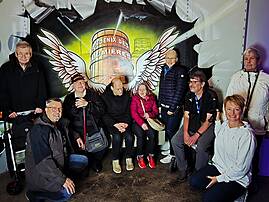 Eine Gruppe von neun Personen posiert lächelnd vor einem beleuchteten Graffiti-Wandbild mit Flügelmotiv und dem Schriftzug „Phoenix des Lumières" in einem abgedunkelten Raum.
