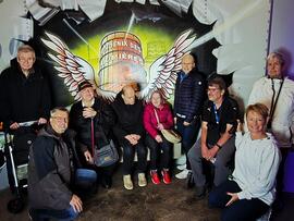  Eine Gruppe von neun Personen posiert lächelnd vor einem beleuchteten Graffiti-Wandbild mit Flügelmotiv und dem Schriftzug „Phoenix des Lumières" in einem abgedunkelten Raum. 
