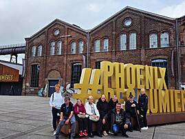 Eine Gruppe von neun Personen sitzt und steht vor großen goldenen 3D-Buchstaben mit dem Schriftzug „#Phoenix des Lumières" auf dem Außengelände eines Backsteingebäudes.