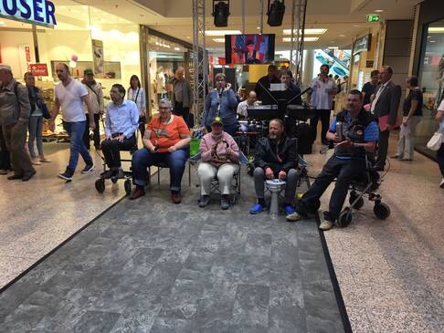 Mehrere Peronen sitzen mit und ohne Hilfsmittel in einem Kaufhaus und machen Musik.