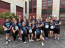 Eine große Gruppe von rund 20 Läuferinnen und Läufern in dunkelblauen „Never Walk Alone"-Shirts posiert lächelnd vor einem Gebäude – viele zeigen Peace-Zeichen oder winken in die Kamera. Es ist das Startfoto des Lebenshilfe-Teams beim Firmenlauf 2026 in Lüdenscheid.