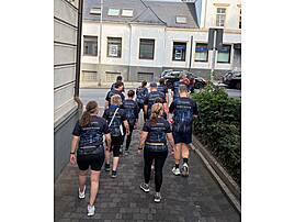 Eine Gruppe von Läuferinnen und Läufern in dunkelblauen Lebenshilfe-Shirts mit dem Aufdruck „Gemeinsam stark. Auf jeder Strecke." läuft von hinten zu sehen eine gepflasterte Straße entlang in Richtung Innenstadt. Das Bild zeigt den Aufbruch des Teams zum Start des Firmenlaufs.