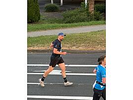 Ein Mann mit blauer Schirmmütze und dunkelblauem Laufshirt läuft konzentriert auf einer asphaltierten Straße an einem Zebrastreifen entlang. Im Hintergrund ist eine Grünanlage zu sehen.
