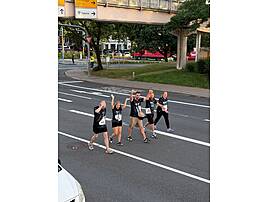 Fünf Läuferinnen und Läufer in dunkelblauen „Never Walk Alone"-Shirts überqueren jubelnd eine Kreuzung in einer Stadt. Sie recken die Arme in die Höhe und feiern gemeinsam.