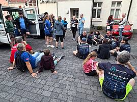 Eine größere Gruppe erschöpfter und erholter Läuferinnen und Läufer versammelt sich nach dem Lauf auf einem Pflasterplatz – einige sitzen auf dem Boden, andere stehen im Gespräch zusammen. Mehrere Personen tragen Shirts der Lebenshilfe Lüdenscheid – Märkischer Kreis mit dem Aufdruck „Gemeinsam stark. Auf jeder Strecke."