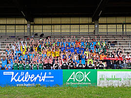 Rund 100 Fußballspielerinnen und -spieler verschiedener Teams stehen jubelnd mit erhobenen Armen auf der Tribüne eines Stadions. Im Vordergrund sind Werbebanden mit den Logos von Küberit und AOK zu sehen.
