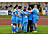 Eine Fußballmannschaft in hellblauen Trikots steht eng zusammen auf dem Rasen und bildet einen Kreis mit den Armen umeinander. Im Hintergrund ist das Stadion mit einer Tartanbahn zu erkennen.