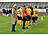 Eine Gruppe von Fußballspielerinnen und -spielern in gelb-schwarzen Trikots mit der Aufschrift „Lüdenscheid-Nord" steht auf dem Spielfeld zusammen. Ein älterer Herr im beigen Mantel steht abseits und beobachtet die Gruppe.