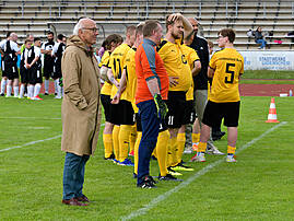 Eine Gruppe von Fußballspielerinnen und -spielern in gelb-schwarzen Trikots mit der Aufschrift „Lüdenscheid-Nord" steht auf dem Spielfeld zusammen. Ein älterer Herr im beigen Mantel steht abseits und beobachtet die Gruppe.
