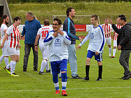 Ein lächelnder Fußballspieler in weißem Trikot mit blauem „Impuls Fairpackung"-Aufdruck steht im Vordergrund auf dem Rasen. Im Hintergrund begrüßen sich Spielerinnen und Spieler verschiedener Teams nach dem Spiel.