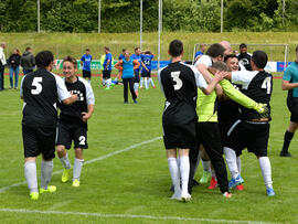 Spielerinnen und Spieler in schwarz-weißen Trikots jubeln auf dem Spielfeld und fallen sich in die Arme. Ein Schiedsrichter im türkisfarbenen Shirt steht am Rand der Szene. 