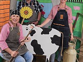 Bild 1: Drei Personen stehen vor einer farbenfrohen, handgemalten Bauernhofkulisse und tragen Schürzen. Gemeinsam präsentieren sie eine große Kuhfigur aus Pappe, während neben ihnen Milchkannen stehen.