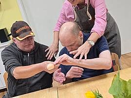 Bild 3: Eine Person mit Kopftuch beugt sich lächelnd über zwei sitzende Personen an einem Tisch. Eine Person hält einen Löffel mit einem Ei. Im Vordergrund stehen Sonnenblumen und bunte Dekoration.