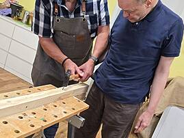 Bild 4: Zwei Männer arbeiten gemeinsam an einer Holzwerkbank; einer trägt einen Strohhut und eine Schürze und hält einen Hammer. Der andere Mann steht daneben und blickt auf das bearbeitete Holzstück.