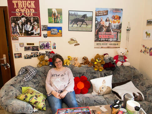 eine Frau sitzt auf dem Sofa auf der Rückenlehne sind Stofftiere und an der Wand im Hintergrund Bilder.