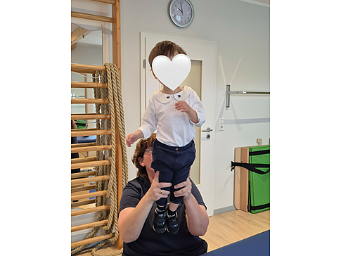 Bild zeigt eine Fachkraft, sie hält ein kleines Kind im Therapieraum hoch.