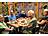 Eine fröhliche Gruppe von fünf Menschen mit Unterstützungsbedarf sitzt gemeinsam an einem Tisch mit Kuchen und Getränken in einem gemütlichen Café mit Pflanzen-Deko.