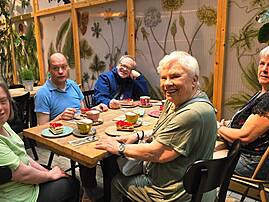 Eine fröhliche Gruppe von fünf Menschen mit Unterstützungsbedarf sitzt gemeinsam an einem Tisch mit Kuchen und Getränken in einem gemütlichen Café mit Pflanzen-Deko.