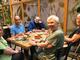 Eine fröhliche Gruppe von fünf Menschen mit Unterstützungsbedarf sitzt gemeinsam an einem Tisch mit Kuchen und Getränken in einem gemütlichen Café mit Pflanzen-Deko. 