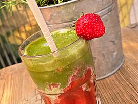 Ein Glas grüner Smoothie oder Limonade mit einer Erdbeere als Deko und einem Strohhalm, daneben eine Schale mit frischen Erdbeeren.