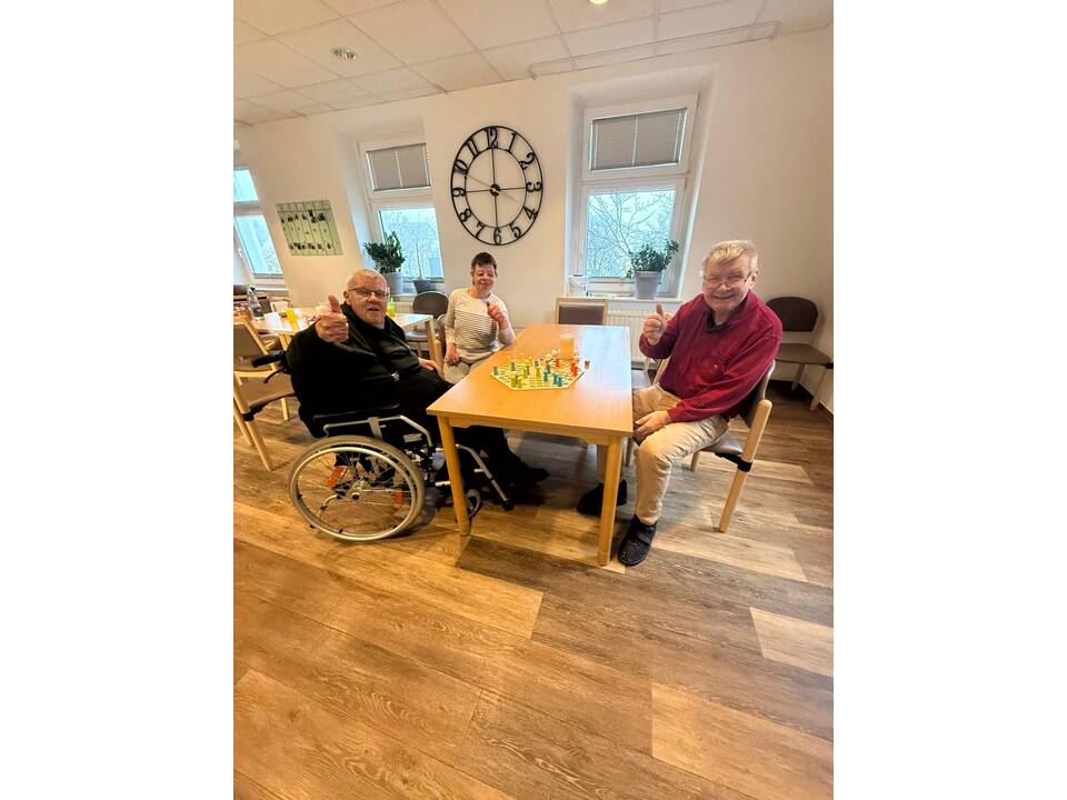 Einige Bewohnerinnen und Bewohner der Wohnstätte Hotopstraße sitzen an einem Tisch und spielen ein Brettspiel. Alle schauen in die Kamera und halten einen Daumen hoch. 