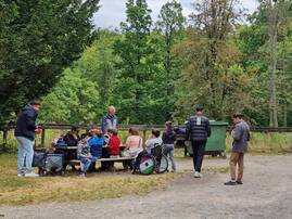 Eine Gruppe von Kindern, Jugendlichen und Erwachsenen macht eine Pause an einem Holztisch mitten im Wald. Unter ihnen ist auch eine Person im Rollstuhl, was die Inklusivität der Gruppe unterstreicht.