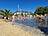 An einem sonnigen Tag vergnügen sich zahlreiche Kinder und Erwachsene auf einem Wasserspielplatz. Sie spielen zwischen Fontänen auf einer dunklen, nassen Fläche, die von Sand und Palmen umgeben ist.