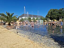An einem sonnigen Tag vergnügen sich zahlreiche Kinder und Erwachsene auf einem Wasserspielplatz. Sie spielen zwischen Fontänen auf einer dunklen, nassen Fläche, die von Sand und Palmen umgeben ist.