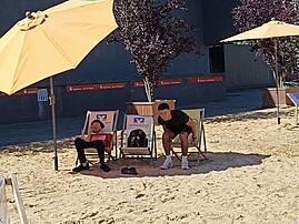 Zwei junge Männer entspannen in Liegestühlen auf einer Sandfläche unter Sonnenschirmen. Im Hintergrund sind Werbebanner der Sparkasse zu sehen.