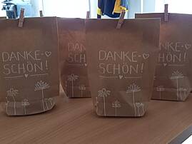 Auf einem Tisch stehen mehrere braune Papiertüten, die mit dem Schriftzug "DANKE SCHÖN!" verziert sind. Jede Tüte ist zusätzlich mit kleinen Herzen und stilisierten Blumen bemalt.