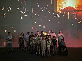 Eine Gruppe Jugendlicher und Erwachsener, darunter eine Person im Rollstuhl, steht für ein Gruppenfoto zusammen. Sie befinden sich in einem abgedunkelten Raum vor einer großen Leinwand mit einer leuchtenden, abstrakten Projektion.