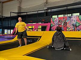 Zwei junge Männer befinden sich in einer Trampolinhalle mit bunten Wänden. Einer von ihnen steht lächelnd auf einer gelben Trampolinfläche, während der andere im Vordergrund sitzt.
