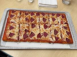 Auf einem metallenen Tablett wird eine große, rechteckige Pizza präsentiert, die in viele Stücke geschnitten ist.