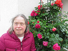  Eine Frau mit rosa Jacke steht neben einem blühenden Rosenstrauch und blickt in die Kamera.