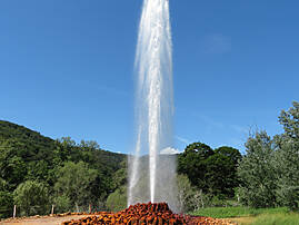 Ein hoher Wasserstrahl schießt vor blauem Himmel und grünem Wald aus einem Brunnen.