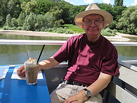 Ein Mann mit Hut sitzt vor einem Fluss in der Sonne und hält ein Getränk