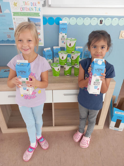 Zwei Mädchen stehen in einem Kindergartenraum und halten jeweils einen Karton Bio-Milch in den Händen. Hinter ihnen sind weitere Milchpackungen und Becher mit Bio-Joghurt zu sehen. Zwei Mädchen stehen in einem Kindergartenraum und halten jeweils einen Karton Bio-Milch in den Händen. Hinter ihnen sind weitere Milchpackungen und Becher mit Bio-Joghurt zu sehen.