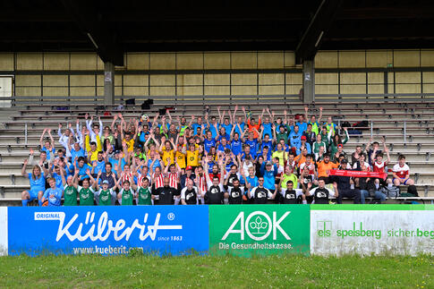 Eine große Gruppe von Menschen in bunten Trikots sitzt auf einer Tribüne in einem Stadion. Sie schauen in die Kamera und winken fröhlich mit den Händen in die Luft. 