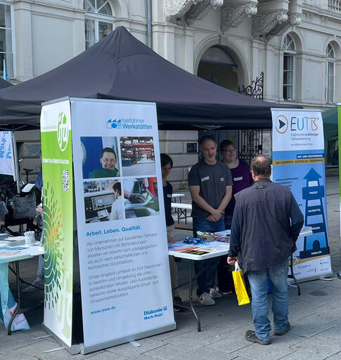 Bild: Infostand unter einem schwarzen Pavillon vor einem historischen Gebäude: Mitarbeitende informieren eine Person am Tisch, daneben stehen Roll-ups der EUTB. Auf dem Tisch liegen Flyer und Materialien.
