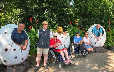 Eine Gruppe von Menschen, darunter Personen im Rollstuhl, posiert gut gelaunt für ein Foto vor einer grünen Hecke. Einige von ihnen sitzen oder stehen in übergroßen, weißen Dinosaurier-Eiern, aus denen sie herausschauen. Eine Gruppe von Menschen, darunter Personen im Rollstuhl, posiert gut gelaunt für ein Foto vor einer grünen Hecke. Einige von ihnen sitzen oder stehen in übergroßen, weißen Dinosaurier-Eiern, aus denen sie herausschauen.