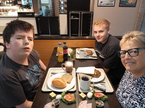 Drei Personen sitzen an einem Frühstückstisch in einem Café. Sie blicken in die Kamera, während sie eine Mahlzeit mit Brötchen, Kaffee und anderen Frühstücksspeisen genießen. Drei Personen sitzen an einem Frühstückstisch in einem Café. Sie blicken in die Kamera, während sie eine Mahlzeit mit Brötchen, Kaffee und anderen Frühstücksspeisen genießen.