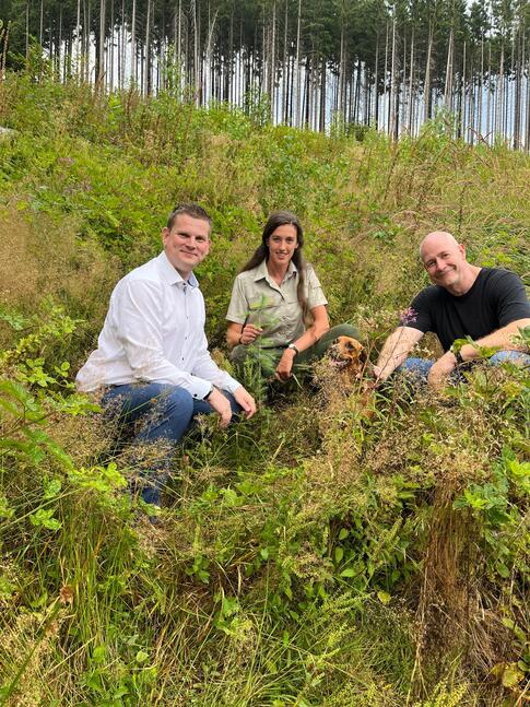Marcel Nüsgen, Geschäftsführer Bürgerstiftung (links), Melanie Fischer von Wald und Holz NRW und Stephan Thiel, Vorstandsvorsitzender Lebenshilfe Lüdenscheid - Märkischer Kreis (rechts) knien mit einem Hund in einer Wiese. Im Hintergrund ist ein durch Trockenheit und Borkenkäfer geschädigter Wald zu erkennen. Frau Fischer zeigt einen frisch gepflanzten Setzling.
