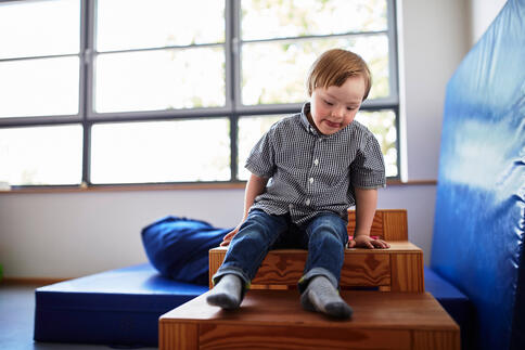 Ein kleiner Junge sitzt konzentriert auf einer Holztreppe in einem hellen Therapieraum. Er trägt ein kariertes Hemd und Jeans und stützt sich mit beiden Händen ab. Ein kleiner Junge sitzt konzentriert auf einer Holztreppe in einem hellen Therapieraum. Er trägt ein kariertes Hemd und Jeans und stützt sich mit beiden Händen ab.
